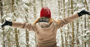 Eine junge Frau im beigen Mantel und einer roten Jacke steht mit ausgebreiteten Armen inmitten eines verschneiten Waldes. - Copyright: Tim Gouw; Pexels